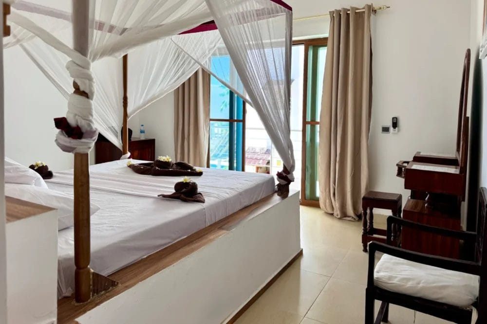 Una luminosa camera d'albergo del BLU MARLIN BEACH RESORT con letto a baldacchino, lenzuola bianche e asciugamani artistici. Le tende chiare coprono le grandi porte di vetro che conducono al balcone, mentre vicino al letto si trovano una scrivania e una sedia in legno.