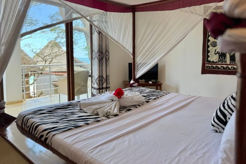 L'accogliente camera da letto del BLU MARLIN BEACH RESORT è dotata di un copriletto zebrato, di un asciugamano con un fiore rosso, di una piccola TV e di ampie finestre che si affacciano su un balcone con vista panoramica sugli alberi, sull'acqua e sulle montagne lontane.