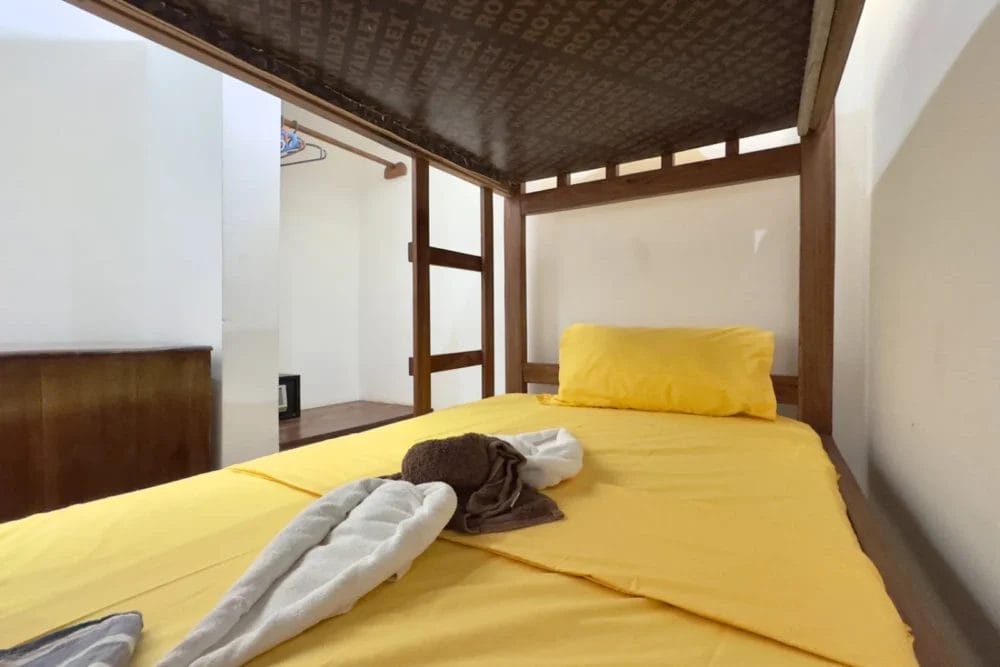 Un letto a castello singolo con lenzuola e cuscino gialli al BLU MARLIN BEACH RESORT. Un asciugamano piegato a forma di cigno si trova sul letto, circondato da pareti bianche semplici e cornici in legno, che creano un'accogliente atmosfera da resort.