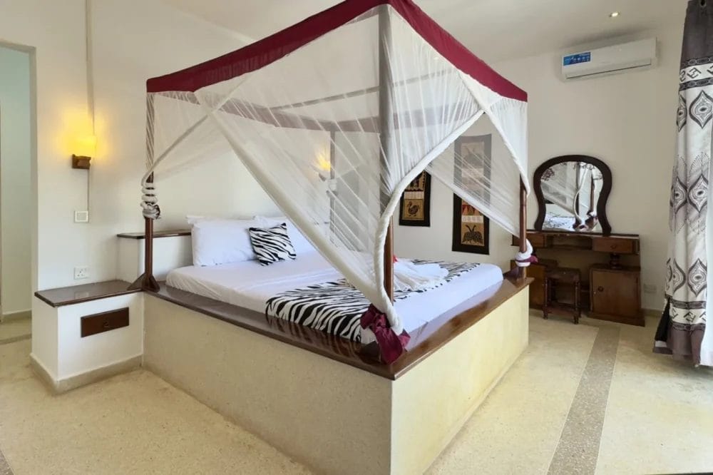 La camera da letto del BLU MARLIN BEACH RESORT, spaziosa e illuminata dal sole, presenta un letto a baldacchino avvolto in una zanzariera bianca, cuscini zebrati, un vanity in legno con specchio e tende a fantasia vicino alla finestra. Le pareti e i pavimenti color crema contribuiscono a creare un'atmosfera ariosa.