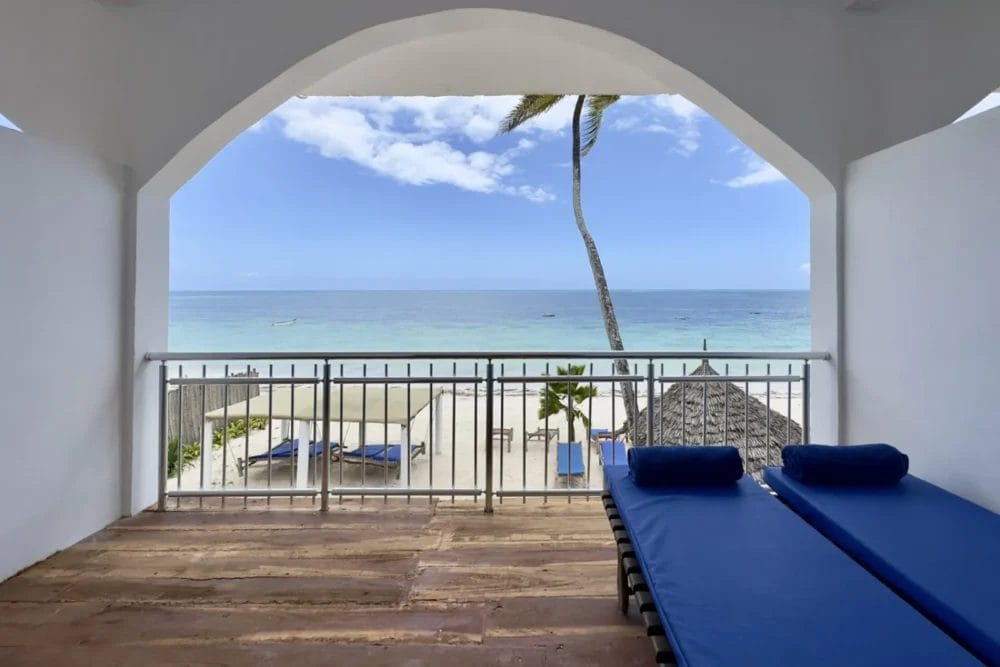 Una terrazza coperta con due sedie a sdraio blu si affaccia sulla spiaggia sabbiosa e sulle palme del BLU MARLIN BEACH RESORT, offrendo una splendida vista sull'oceano calmo e blu sotto un cielo parzialmente nuvoloso.
