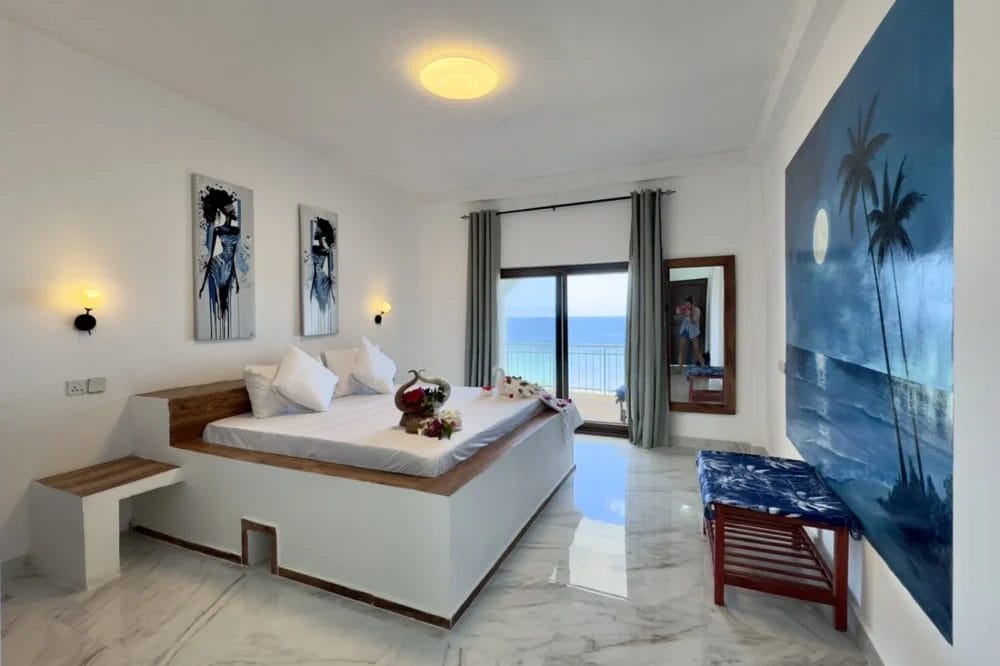 Camera moderna e luminosa del BLU MARLIN BEACH RESORT con letto grande, balcone con vista sull'oceano e pavimenti in marmo. Opere d'arte a tema tropicale, due lampade a muro, una panca e tende grigie incorniciano la splendida vista sul mare di questo resort sulla spiaggia.