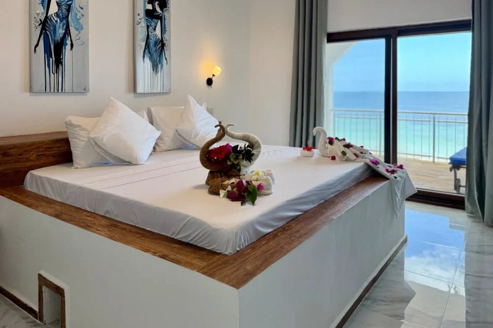Un letto ben fatto con lenzuola bianche, cuscini e sculture di asciugamani - cigno ed elefante - decorati con fiori. Una grande finestra rivela l'oceano e il balcone con una sedia a sdraio blu del BLU MARLIN BEACH RESORT.