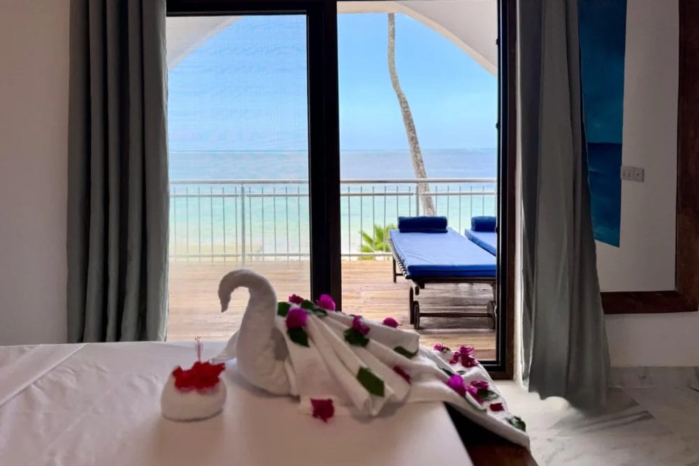 Un asciugamano piegato a forma di cigno con petali di fiori giace sul letto di una camera del BLU MARLIN BEACH RESORT, affacciata su un balcone con sedie a sdraio blu e vista sull'oceano attraverso grandi porte di vetro.