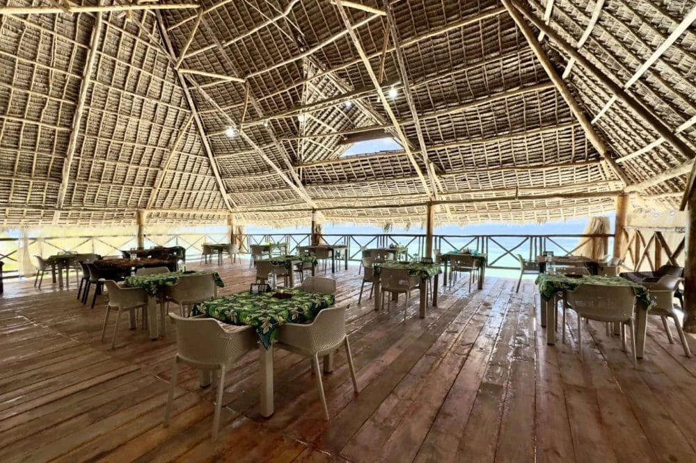 Ampio ristorante all'aperto del BLU MARLIN BEACH RESORT con un alto tetto di paglia, pavimento in legno e tavoli ricoperti di stoffe a motivi verdi. L'area ben illuminata si affaccia su una vista panoramica attraverso ringhiere di legno di questo invitante resort sulla spiaggia.