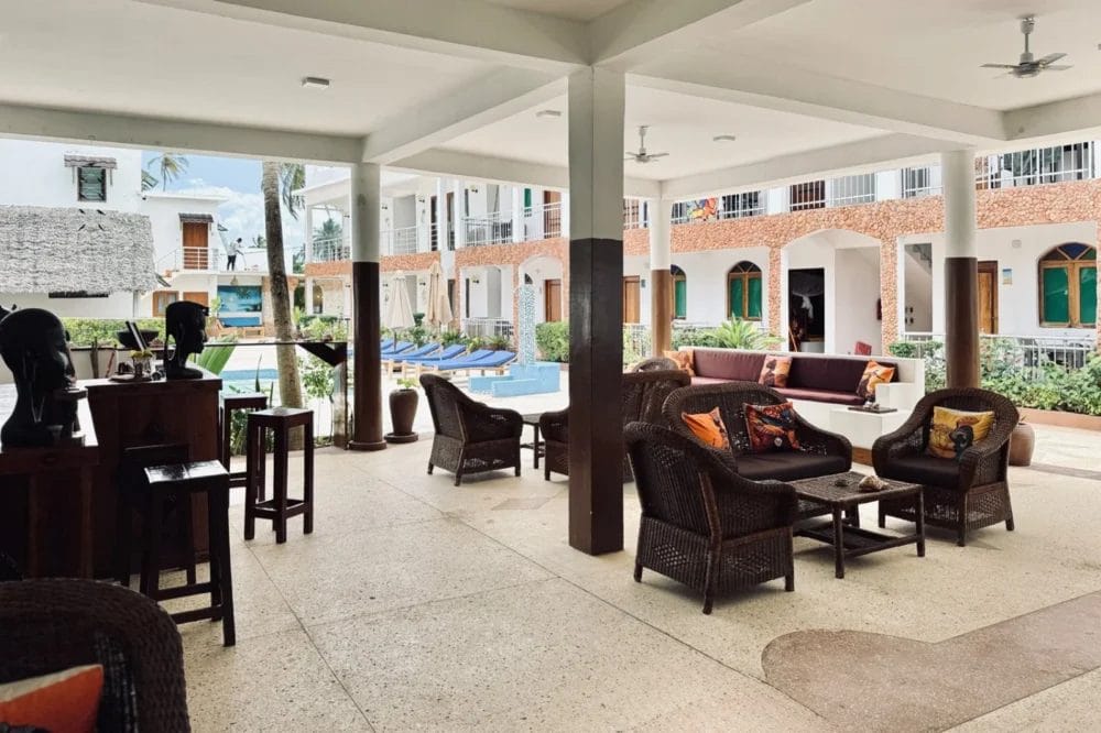 Area lounge coperta all'aperto con sedie e divani in vimini, affacciata su una piscina circondata da lettini. L'edificio a due piani con balconi e piante tropicali sullo sfondo crea un'atmosfera rilassata e da resort BLU MARLIN BEACH RESORT.