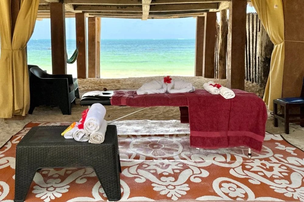 Un lettino per massaggi ricoperto di asciugamani rossi e asciugamani bianchi arrotolati si trova su un tappeto a fantasia in una cabina all'aperto del BLU MARLIN BEACH RESORT, affacciata sull'oceano, con sedie a sdraio e tende che impreziosiscono questo ambiente sereno del resort sulla spiaggia.