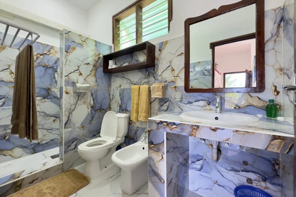 Il bagno moderno del BLU MARLIN BEACH RESORT è caratterizzato da piastrelle marmorizzate blu e oro, wc, bidet, lavandino con specchio, doccia in vetro, portasciugamani con asciugamani beige e una finestra in alto: l'ideale per il vostro rifugio al mare.