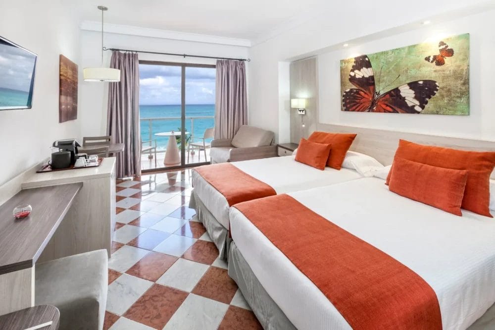Moderna camera d'albergo MELIA VARADERO con due letti, accenti arancioni, opere d'arte di farfalle, piccola scrivania, TV, poltrona e porte scorrevoli in vetro che si aprono su un balcone con tavolo, sedie e splendida vista sull'oceano in questo resort di Cuba.