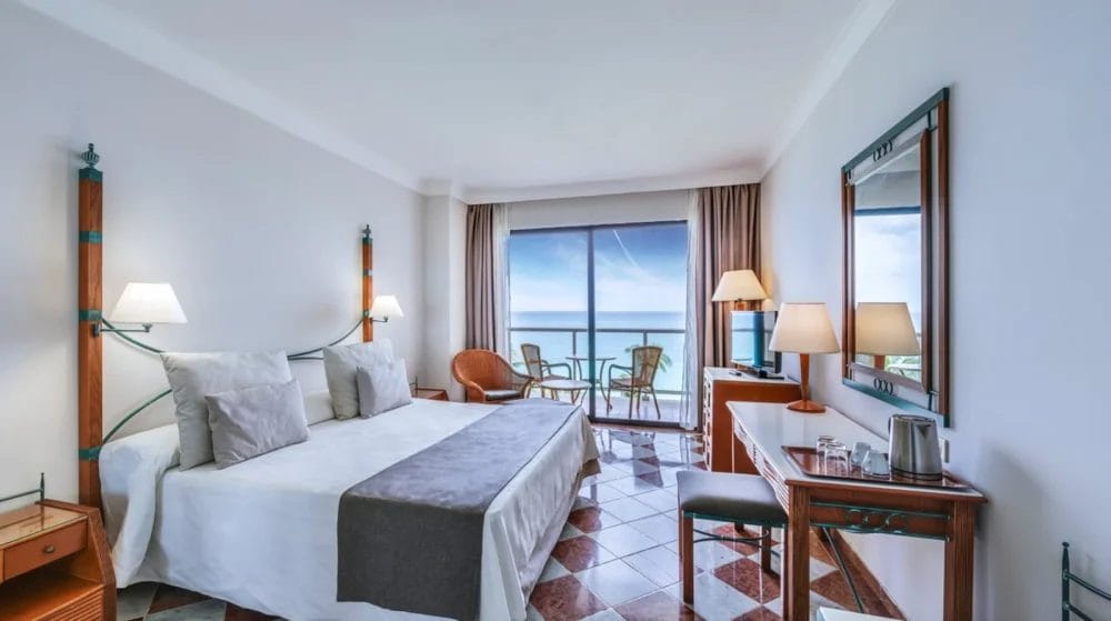 Una luminosa camera d'albergo di MELIA VARADERO con un grande letto, tavolini e lampade. Sulla destra si trova una scrivania con specchio, bollitore e tazze. In questa destinazione di MELIA Cuba c'è un balcone con sedie e una splendida vista sull'oceano.