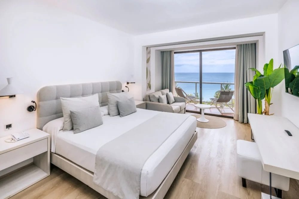 Una moderna camera d'albergo a Varadero con un grande letto, arredi grigi e bianchi, divano, scrivania e porte scorrevoli in vetro che conducono a un balcone con vista sull'oceano e sulle palme di questo splendido resort di Cuba.