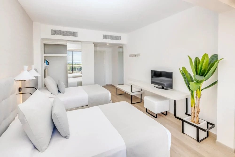 Una luminosa e moderna camera dell'hotel Melia Varadero con due letti matrimoniali, pavimento in legno chiaro, TV a schermo piatto su una console bianca, una pianta verde in vaso e ampie finestre con vista sull'esterno. L'arredamento è minimalista e prevalentemente bianco.