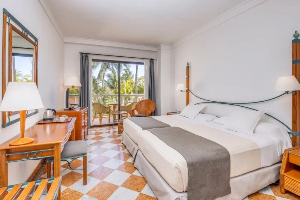 La luminosa camera dell'hotel MELIA di VARADERO è dotata di un grande letto, di un pavimento con piastrelle a scacchi, di una scrivania e di una sedia in legno, di sedie in vimini, di una lampada e di porte scorrevoli in vetro che si aprono su un balcone con palme all'esterno.