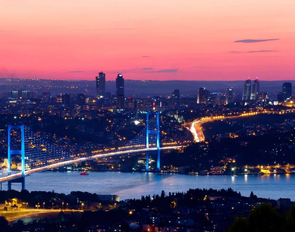 Un ponte sospeso illuminato, che ricorda il PONTE 2 GIUGNO di Istanbul, attraversa un ampio fiume al tramonto, collegando due paesaggi urbani pieni di grattacieli e luci incandescenti sotto un cielo colorato di rosa e arancione.