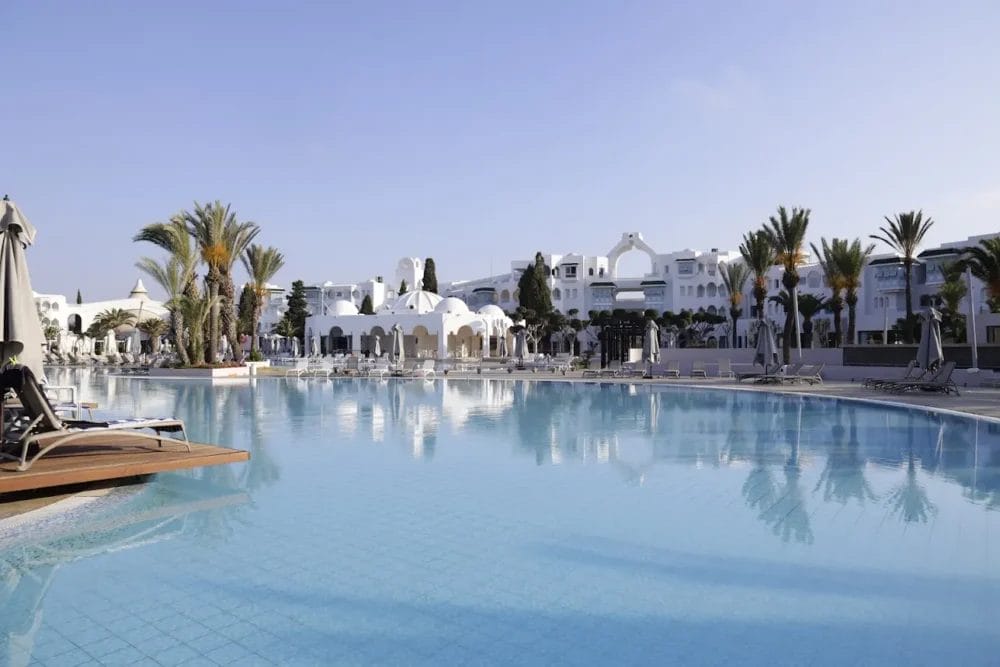 Una grande piscina all'aperto circondata da sedie a sdraio, palme ed edifici bianchi con cupole e archi sotto un cielo azzurro e limpido nel lussuoso Mirage Hammamet Resort, un rinomato hotel a 5 stelle.