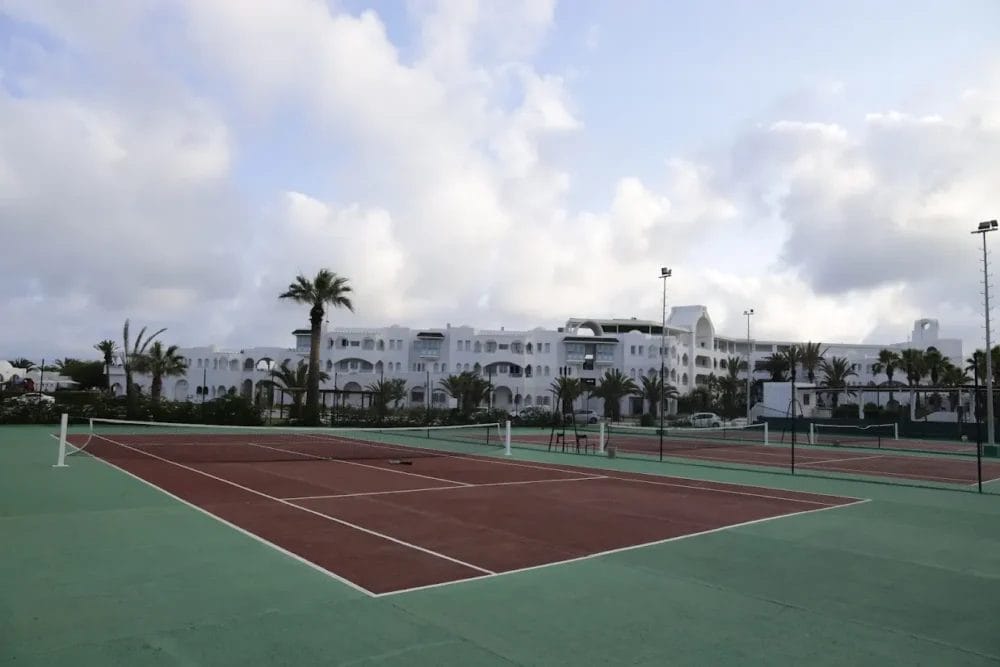 Campi da tennis all'aperto con superficie rossa e ambiente verde, vicino a palme e al grande resort MIRAGE HAMMAMET, caratterizzato da un grande edificio bianco con archi sotto un cielo parzialmente nuvoloso, perfetto per un soggiorno attivo o per il relax post-partita alla SPA.
