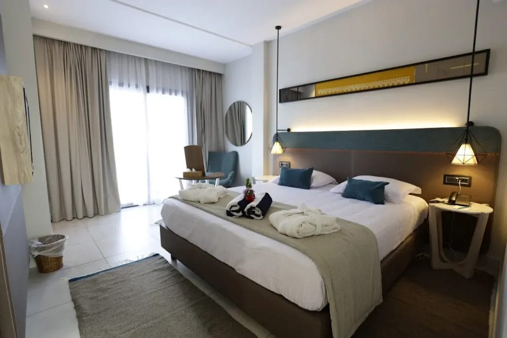 Una moderna camera d'albergo RESORT al MIRAGE HAMMAMET con un grande letto matrimoniale, due accappatoi, cuscini blu d'accento, un tavolino con vino e bicchieri, una poltrona vicino alla finestra con tende e un'illuminazione d'ambiente soffusa.