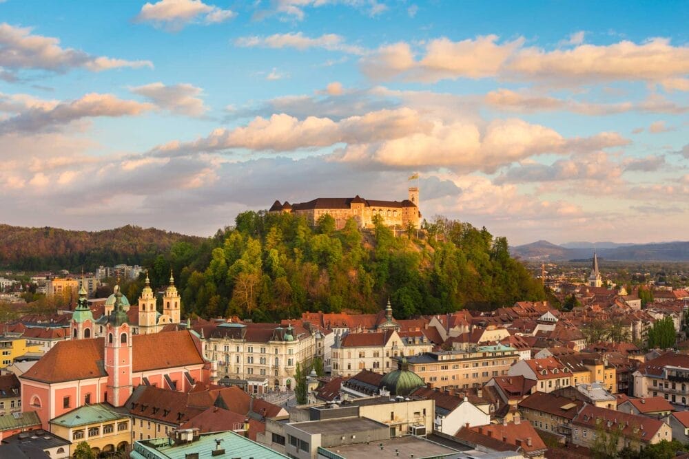 Un castello in cima a una collina in Slovenia, circondato da alberi verdi, domina una città con edifici dai tetti rossi sotto un cielo parzialmente nuvoloso al tramonto: l'ideale per vivere i colori dell'autunno nel vostro prossimo tour granturismo.