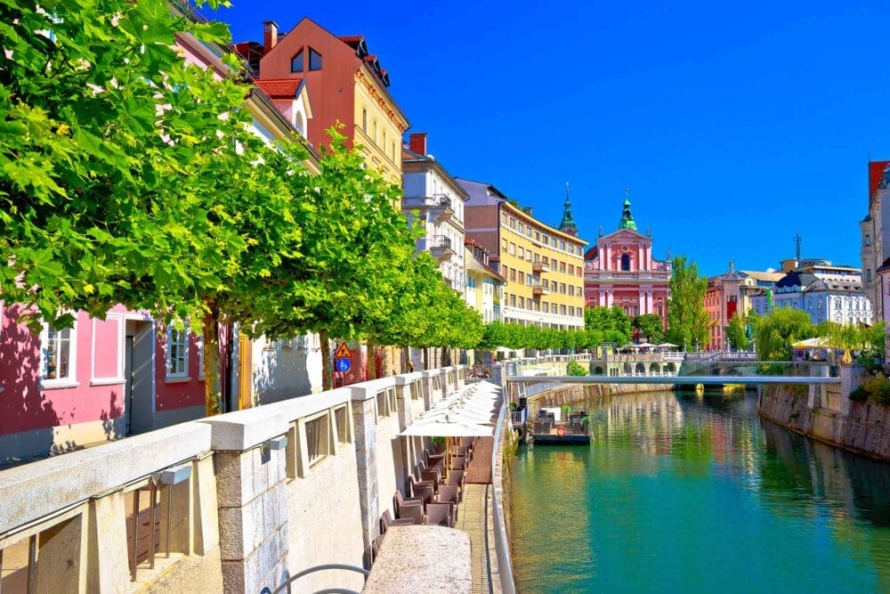 Edifici colorati e alberi frondosi con i colori dell'autunno costeggiano un canale con posti a sedere all'aperto in una soleggiata città europea; una chiesa rosa con cupole verdi si staglia contro il cielo blu brillante, creando una scena affascinante in Slovenia.