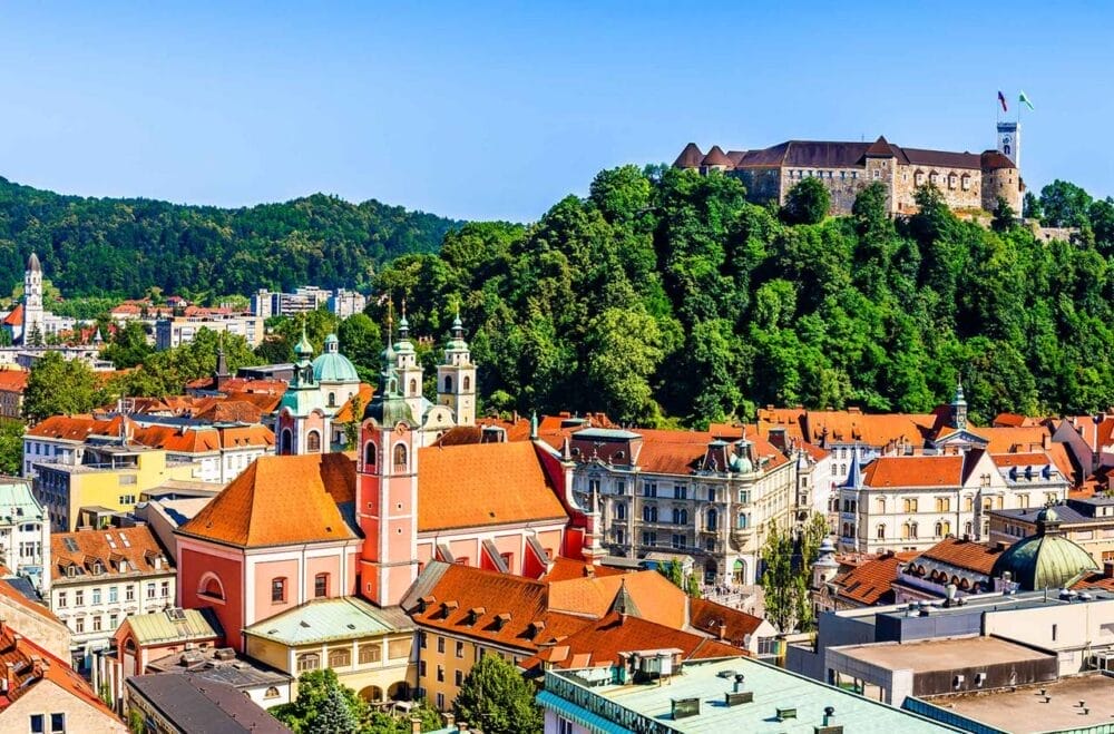 Una vista panoramica di Lubiana, in Slovenia, con edifici dai tetti rossi e una chiesa rosa incastonata sotto il Castello di Lubiana in cima a una collina verde, il tutto illuminato dai vibranti colori dell'autunno sotto un cielo azzurro e limpido.