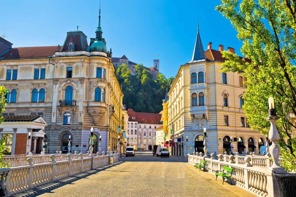 Una scena di strada soleggiata a Lubiana, in Slovenia, è caratterizzata da edifici storici ornati, un ponte di ciottoli e alberi adornati con i colori dell'autunno, mentre il castello di Lubiana si erge su una collina sotto un cielo azzurro e limpido.