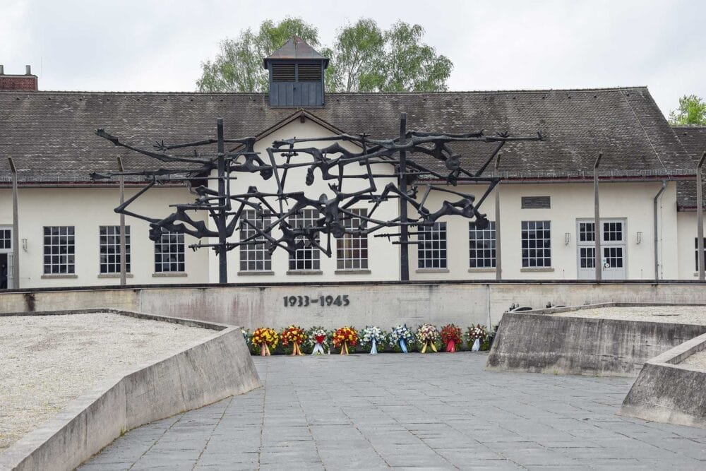 Una scultura commemorativa di figure umane aggrovigliate si trova davanti a un edificio del campo di concentramento di Dachau, vicino a Monaco di Baviera (Monaco), con fiori in basso e l'indicazione degli anni "1933-1945": una tappa fondamentale di qualsiasi tour granturismo in Baviera.