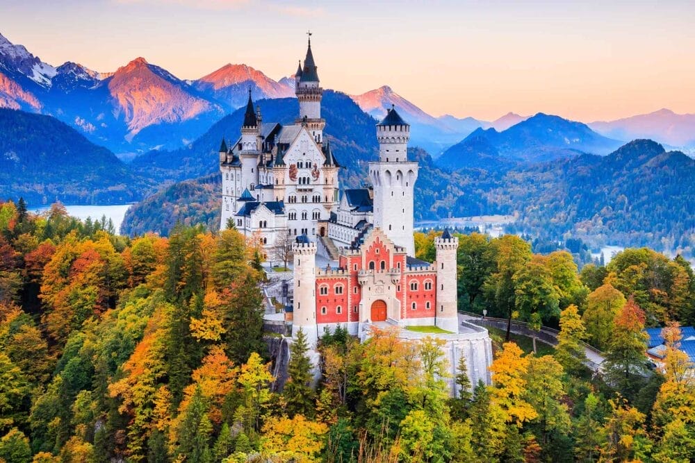 Il Castello di Neuschwanstein, uno dei famosi Castelli della Baviera, si erge in cima a una collina boscosa circondata da alberi autunnali, cime montuose e un lago: una vista incantevole che si gode spesso durante un tour Pullman GranTurismo da Monaco al tramonto.