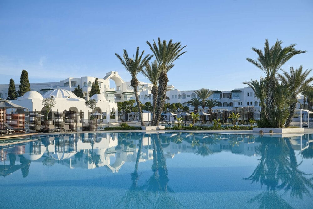 La grande piscina all'aperto del MIRAGE HAMMAMET RESORT & SPA a 5 stelle riflette le palme e gli edifici bianchi in stile mediterraneo sotto un cielo azzurro e limpido, creando una tranquilla atmosfera da resort.
