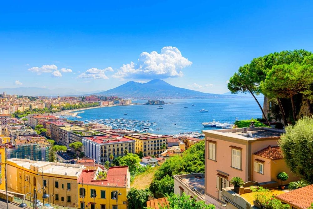 Edifici costieri colorati e un porto turistico costeggiano la riva sotto il cielo azzurro di Napoli, con alberi rigogliosi in primo piano e un grande vulcano coronato da nuvole che si erge sullo sfondo.