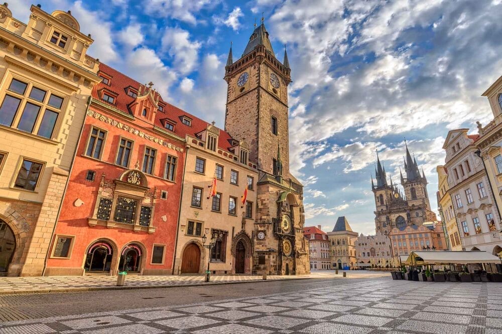 Una piazza storica di Budapest o Praga, con edifici colorati, un'alta torre dell'orologio e una piazza in pietra a motivi sotto un cielo parzialmente nuvoloso. Poche persone sono visibili, il che la rende ideale per un tour in Pullman Granturismo in mezzo all'architettura ornata.
