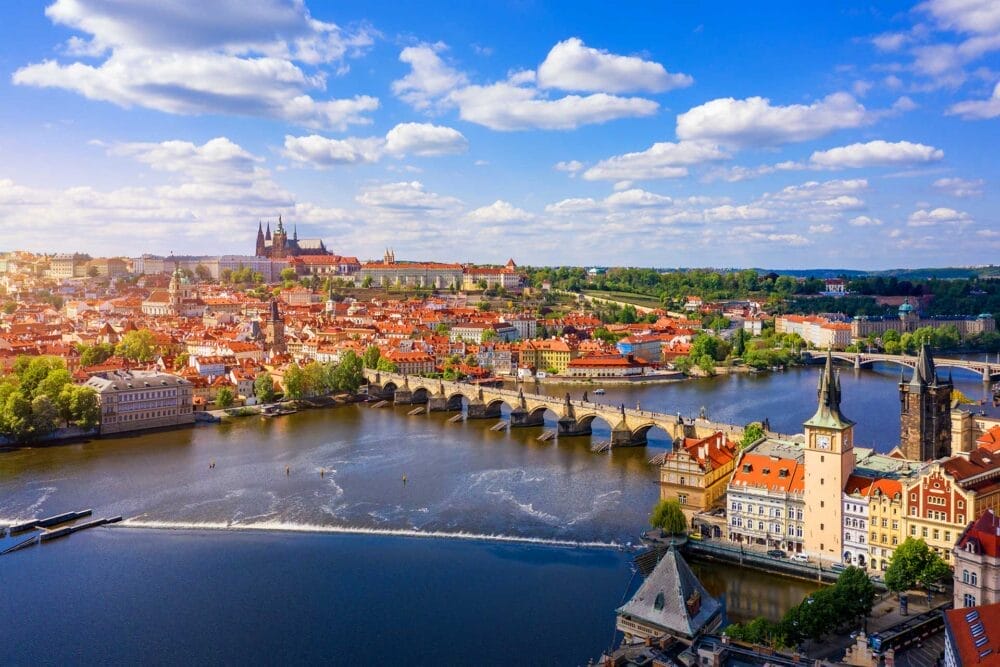 Una vista panoramica di Praga con lo storico Ponte Carlo che attraversa il fiume Moldava, gli edifici dai tetti rossi, la vegetazione lussureggiante e il Castello di Praga in lontananza: l'ideale per un memorabile tour Granturismo sotto un cielo azzurro con nuvole sparse.