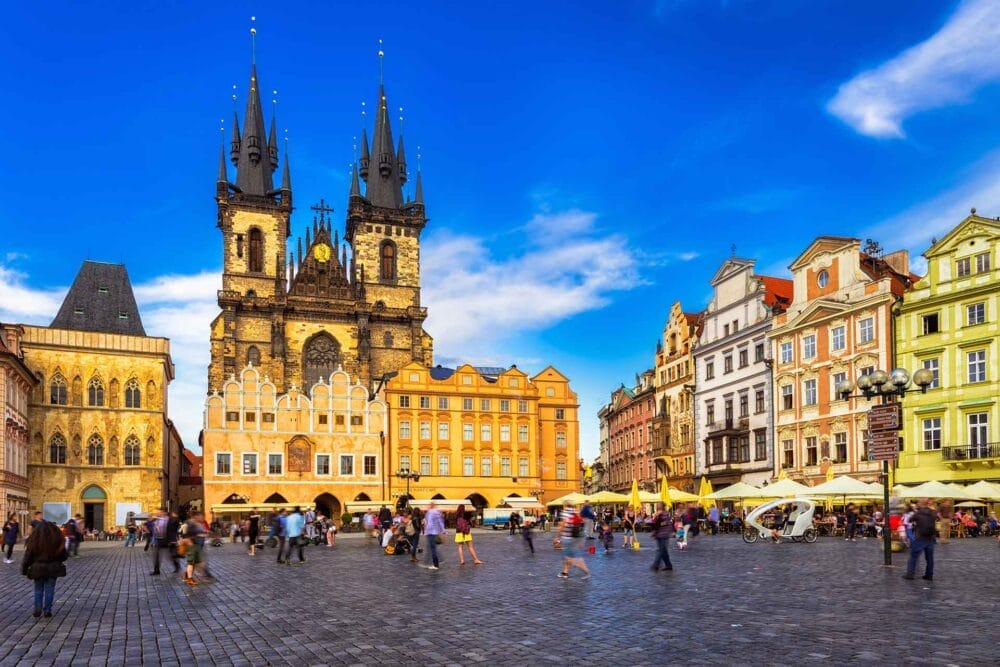 Una vivace piazza della città, come quelle di Praga o Budapest, è caratterizzata da edifici storici colorati, una grande chiesa gotica con alte guglie, persone che si riuniscono e caffè all'aperto, il tutto facilmente esplorabile in un tour Pullman Granturismo sotto un cielo blu brillante.