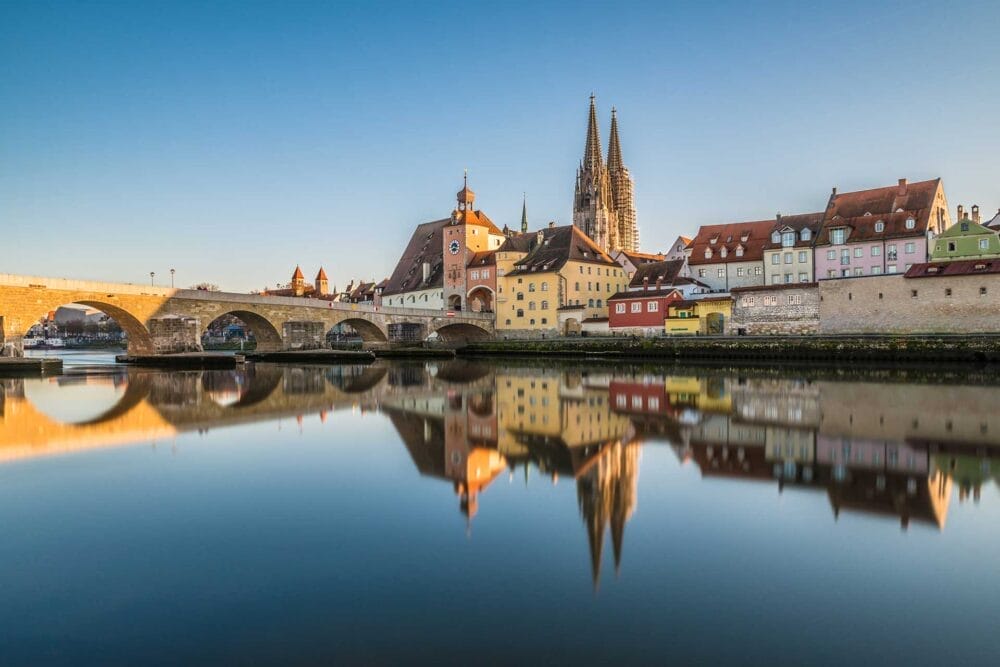 Una vista panoramica di una storica città europea, che ricorda Budapest, con edifici colorati, un ponte in pietra e una grande cattedrale con due guglie, il tutto riflesso in un fiume calmo sotto un cielo azzurro e limpido: l'ideale per qualsiasi tour granturismo.