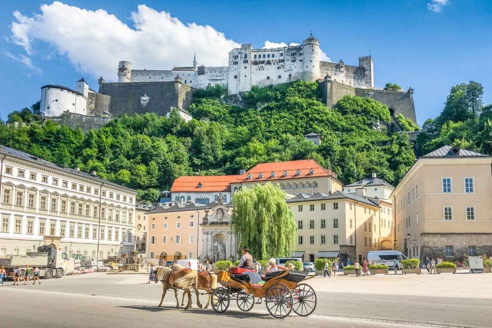 Una carrozza trainata da cavalli attraversa una soleggiata piazza europea di Vienna, con edifici storici colorati e un grande castello in cima a una collina verdeggiante sullo sfondo.