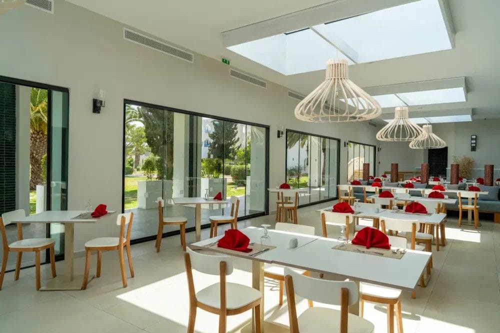 Il ristorante del Mirage Hammamet Resort è luminoso e moderno, con tavoli bianchi e sedie in legno. I tovaglioli rossi sono ordinatamente piegati su ogni tavolo. Grandi finestre e lucernari riempiono lo spazio dell'hotel a 5 stelle di luce naturale, completata da luci a sospensione minimaliste.
