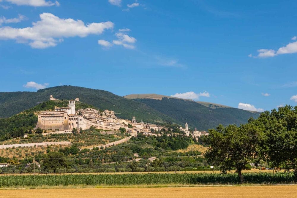 Una città collinare con edifici storici in pietra e torri si trova sotto montagne verdi, con campi, alberi e un cielo azzurro - catturando il fascino senza tempo dell'Umbria classica per qualsiasi itinerario di viaggio da sogno in Umbria.