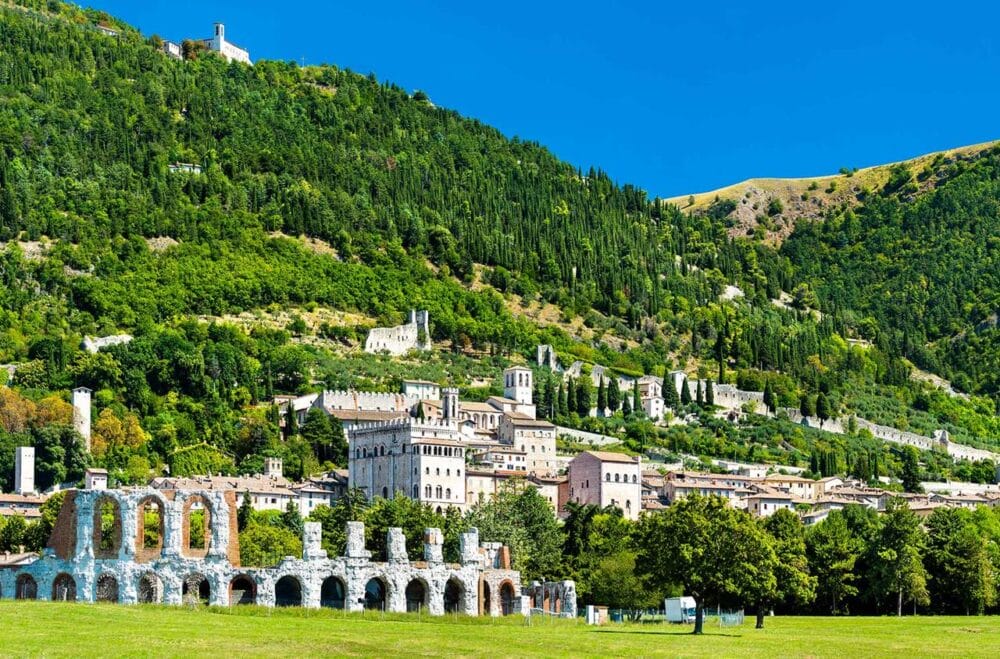 Una vista panoramica di una città collinare italiana vi attende durante la vostra avventura in Umbria Classica, con una vegetazione lussureggiante, edifici storici in pietra, antiche rovine in primo piano e un cielo azzurro e limpido che fa da sfondo perfetto.
