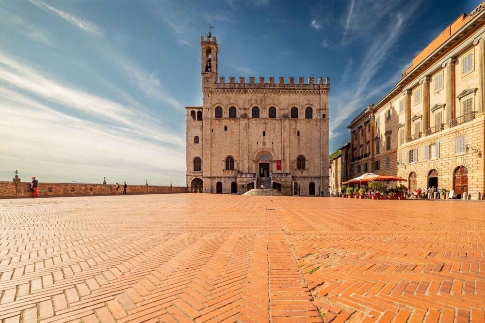 Una piazza ampia e vuota, lastricata di mattoni, si affaccia su un edificio storico in pietra con finestre ad arco e una torre dell'orologio sotto un cielo azzurro: una scena classica che incontrerete durante il vostro TOUR UMBRIA CLASSICA, con persone che si rilassano in un caffè sotto gli ombrelloni nelle vicinanze.