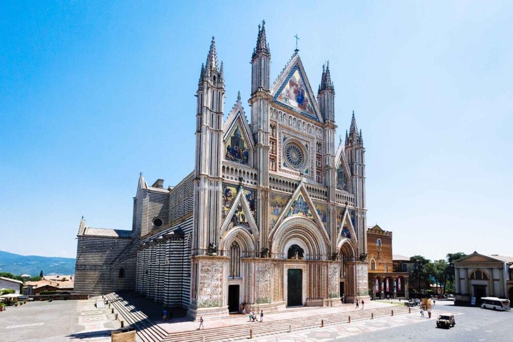 L'immagine mostra l'ornata facciata del Duomo di Orvieto, in Italia, uno dei punti di forza del viaggio in Umbria, caratterizzato da architettura gotica, mosaici colorati, alte guglie e intricate sculture in un cielo azzurro e una piazza aperta.