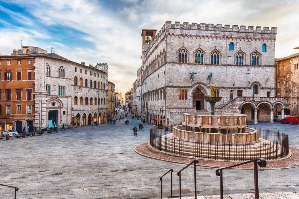 Un'ampia piazza in pietra di fronte a edifici storici, con una grande fontana decorata e statue in primo piano. Le persone camminano lungo la piazza aperta sotto un cielo parzialmente nuvoloso: un luogo perfetto per vivere l'Umbria classica durante il vostro tour dell'Umbria.
