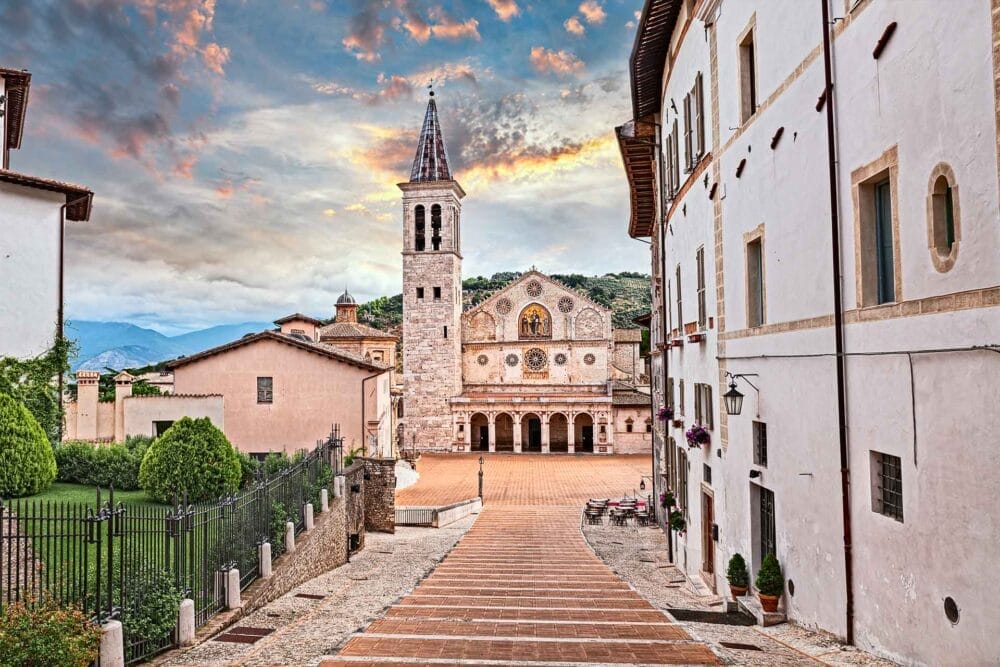 Una strada lastricata in pietra conduce a una chiesa ornata con un alto campanile, una facciata intricata, archi e rosoni - una scena iconica dell'Umbria classica circondata da edifici storici sotto un cielo colorato dal tramonto.