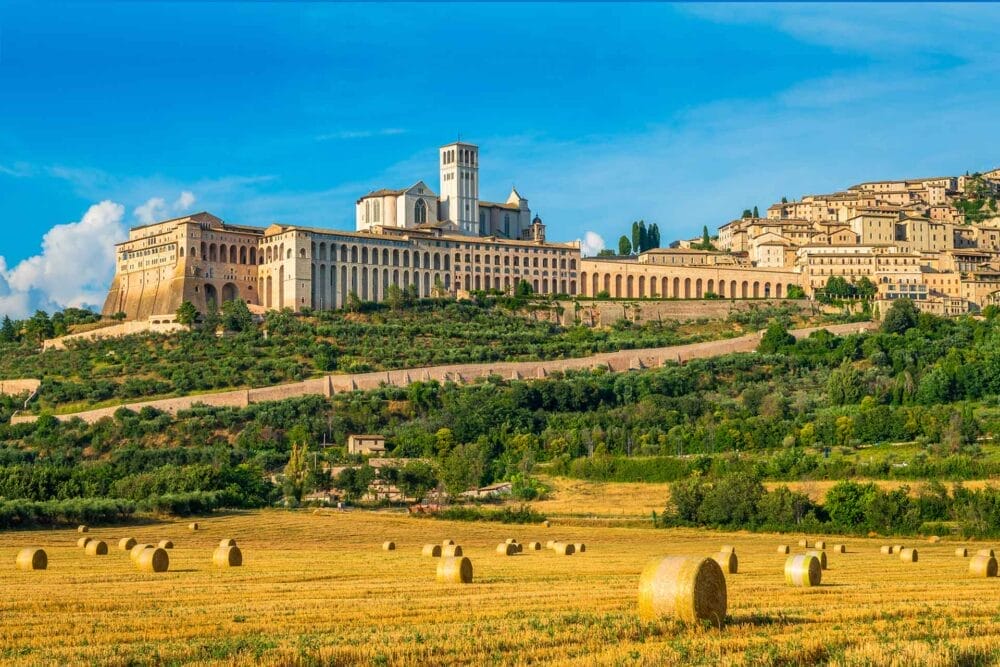 Una vista panoramica della Basilica di San Francesco sulla cima di una collina ad Assisi, in Italia - un punto culminante di qualsiasi tour classico - circondata da edifici abbronzati, verde lussureggiante e balle di fieno sotto il cielo blu brillante dell'Umbria.