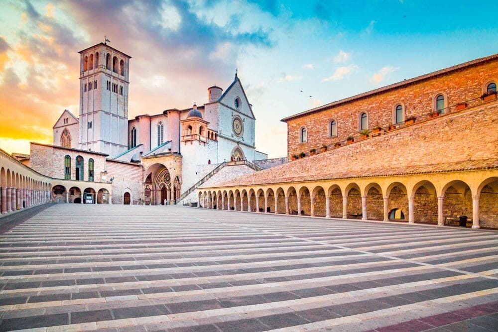 Un'ampia piazza in pietra a strisce conduce alla Basilica di San Francesco d'Assisi in Umbria, caratterizzata da camminamenti ad arco, un alto campanile e un cielo spettacolare all'alba o al tramonto: un punto culminante di qualsiasi tour classico del turismo umbro.