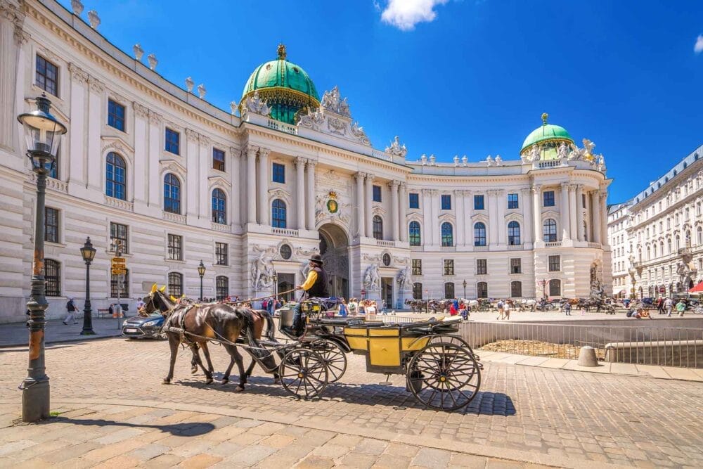 Una carrozza trainata da cavalli passa davanti all'imponente ingresso a cupola del Palazzo Hofburg di Vienna, in Austria, sotto un cielo azzurro e limpido, mentre le persone che passeggiano e si rilassano nelle vicinanze assorbono il fascino della loro esperienza di Tour Austria.