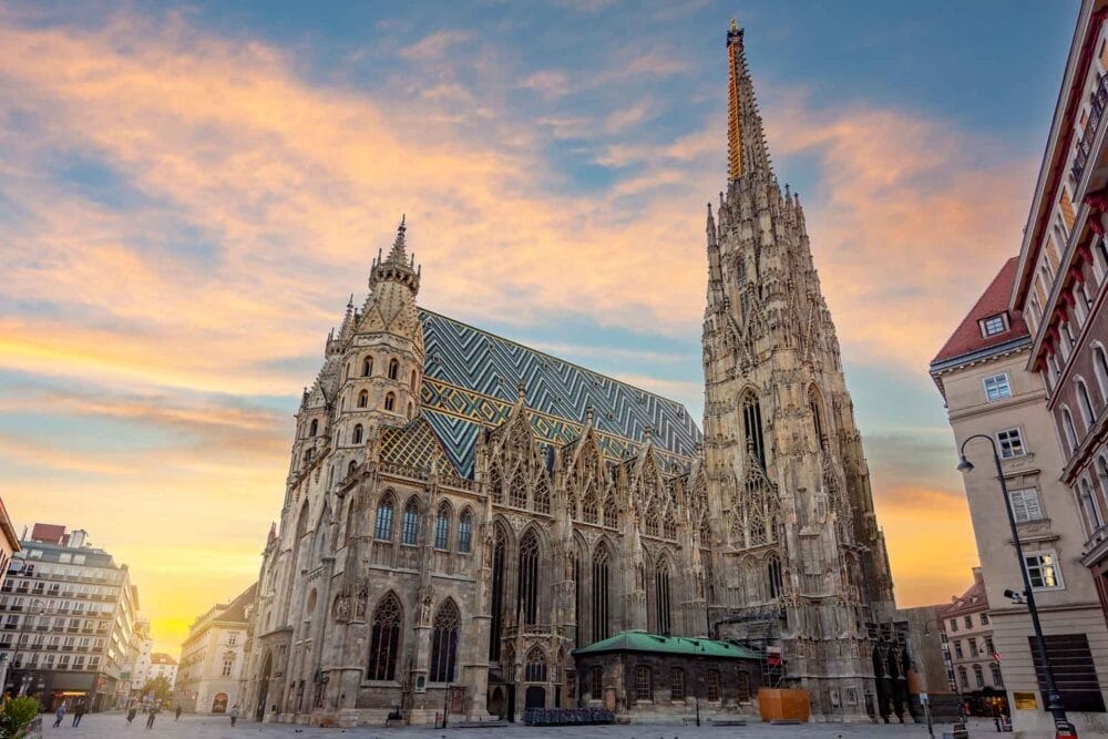 Una grande cattedrale gotica con un tetto di tegole decorate e alte guglie si erge in una piazza della città al tramonto, circondata da edifici storici: una vista iconica in qualsiasi Tour dell'Austria, specialmente quando si visita Vienna.