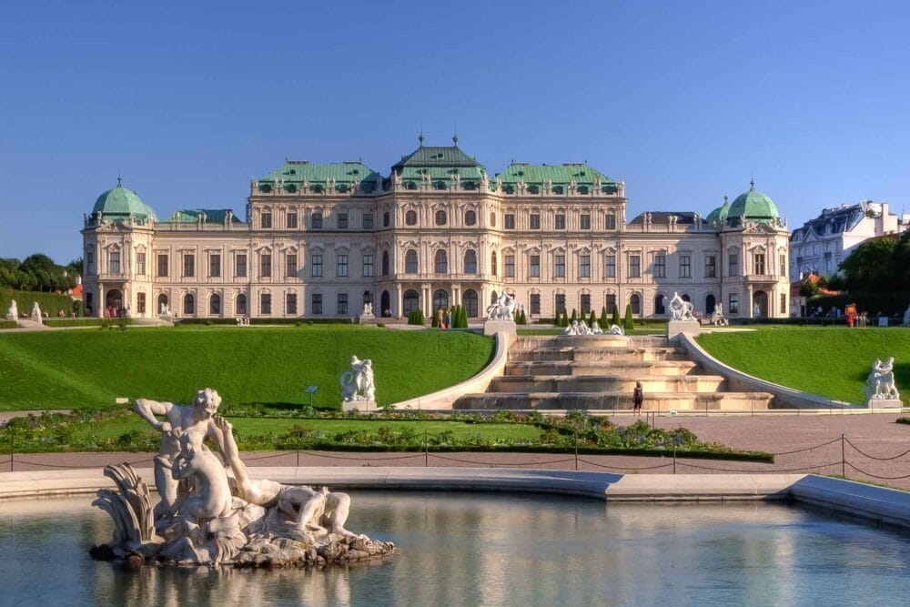 Un grandioso palazzo barocco con cupole verdi e statue ornate si erge dietro giardini curati e una fontana decorativa, sotto il cielo azzurro e limpido di Vienna: un'occasione imperdibile per visitare l'Austria.