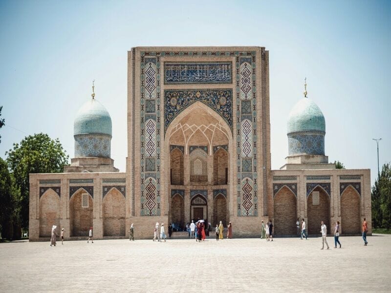 La gente cammina davanti a una grande moschea in mattoni decorata con due cupole turchesi e intricate piastrelle sulla facciata: una vista iconica in un tour classico dell'Uzbekistan sotto un cielo azzurro e limpido.