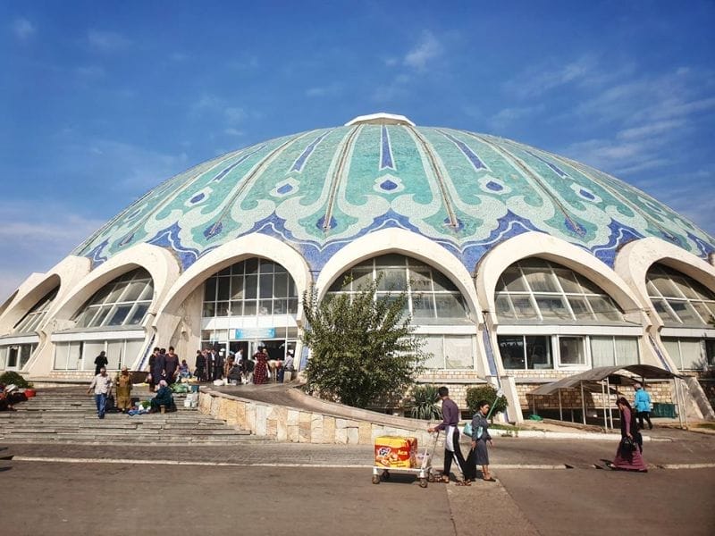 Un grande edificio a cupola con piastrelle a mosaico blu e verdi, finestre ad arco e persone riunite all'esterno vicino alle bancarelle del mercato sotto un cielo azzurro e limpido: una tappa iconica del tour della Carovana in Uzbekistan, prima di dirigersi verso il campeggio yurta sul lago Aydarkul.