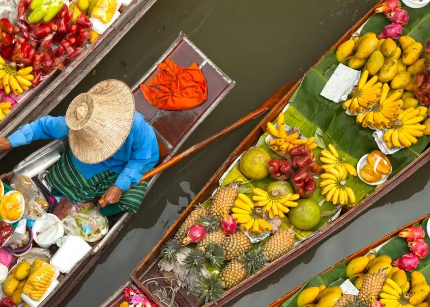Vista aerea di barche su un fiume in un mercato galleggiante di Phuket, pieno di frutta colorata come banane, ananas e mango. Una persona con cappello di paglia e camicia blu siede su una barca, circondata da prodotti durante un Tour Tailandia.