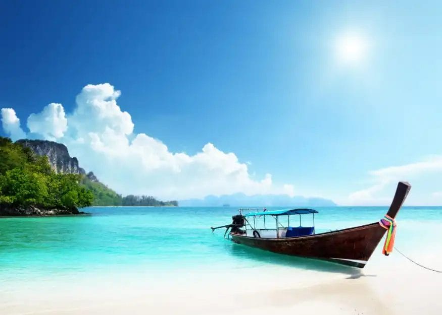 Una tradizionale barca a coda lunga in legno galleggia sull'acqua limpida e turchese vicino a una spiaggia sabbiosa di Phuket, con scogliere verdeggianti e un cielo azzurro brillante: una scena iconica che attende chi fa il Tour Thailandia di prestigio.